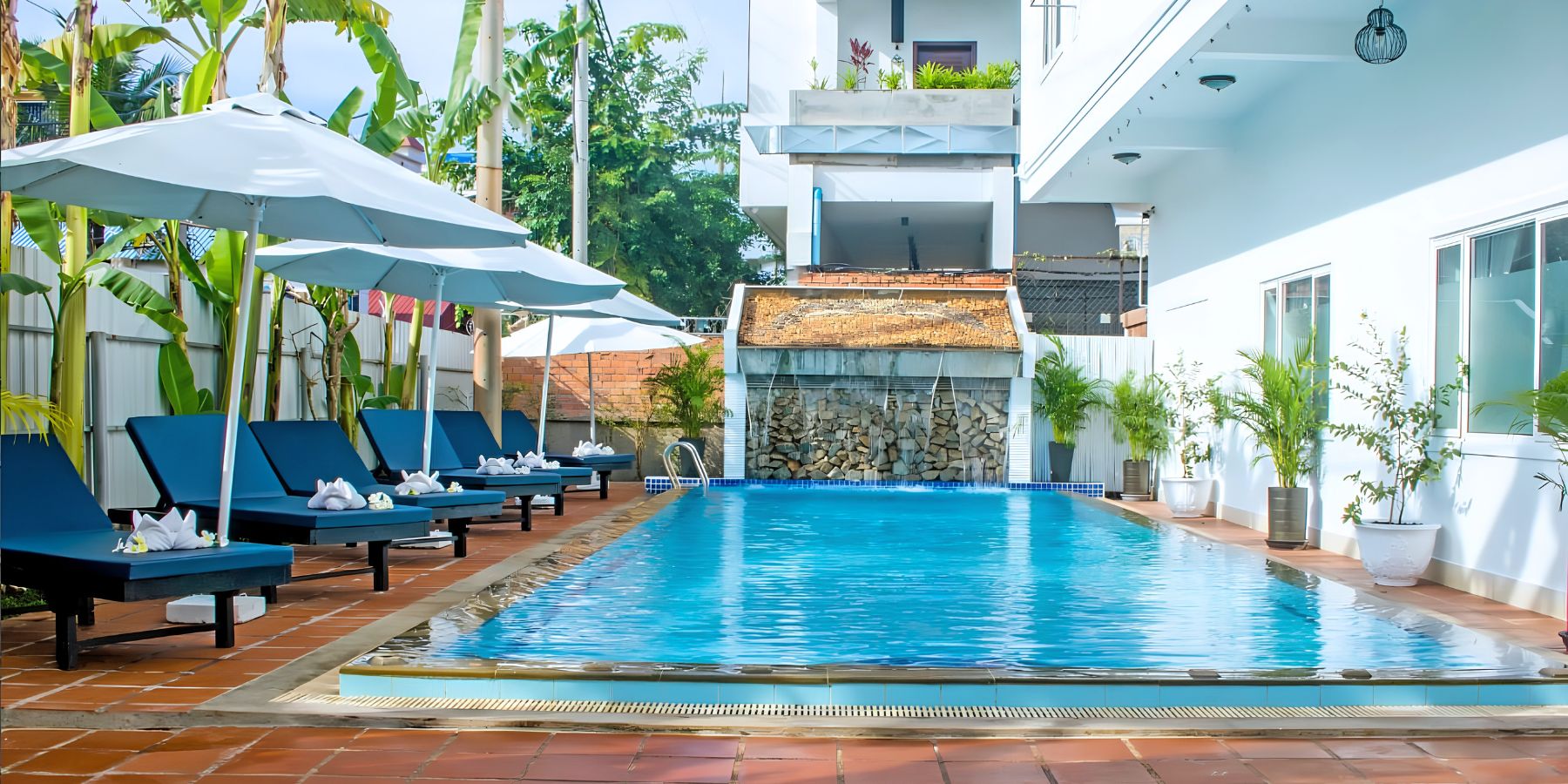 Long outdoor resort swimming pool surrounded by blue sun loungers, white umbrellas, and a waterfall wall.