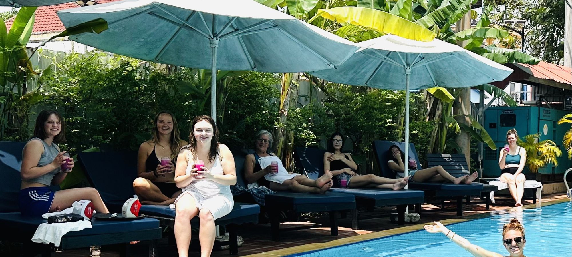 Group of tourists relaxing on blue sun loungers holding tropical drinks by a resort pool.