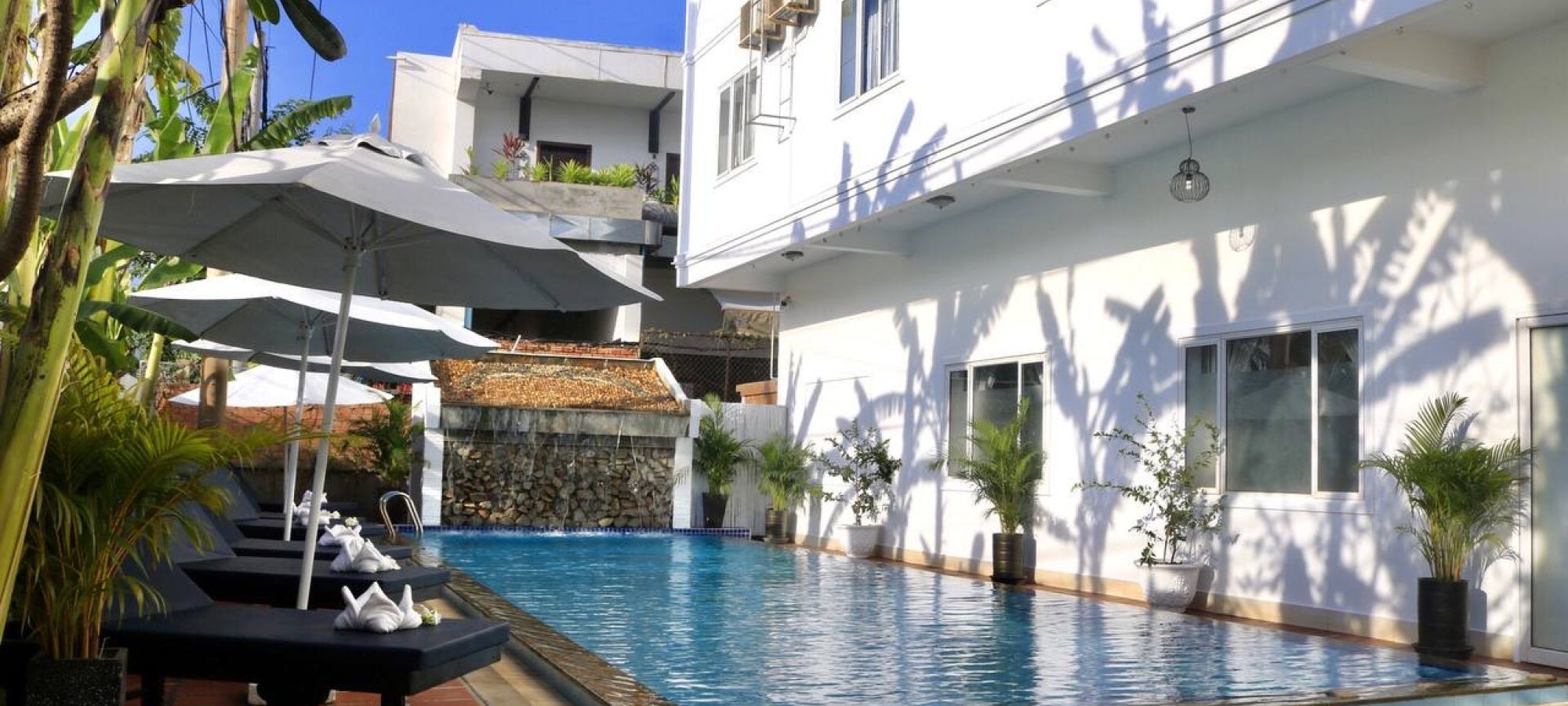 Outdoor swimming pool alongside a white hotel building lined with lounge chairs and shade umbrellas.
