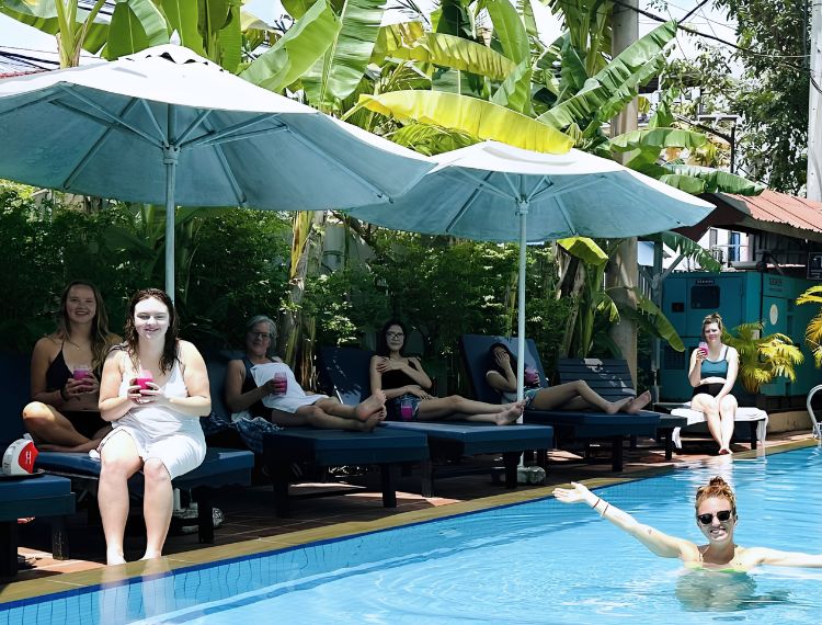 Guests relaxing on poolside sun loungers under white patio umbrellas at a tropical resort pool.