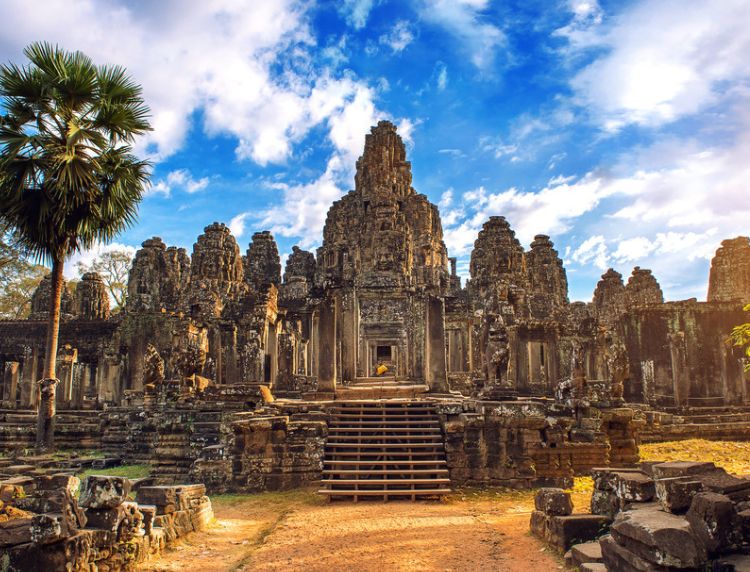 Stone face towers of Bayon Temple in Angkor Thom framed by a palm tree under a blue sky with soft clouds.
