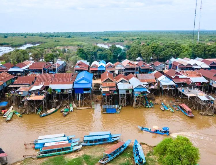 Aerial view of a floating village with colorful stilted houses along a brown river and green forest in Tonle Sap, Cambodia.