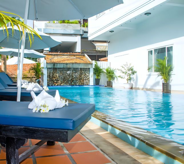 Resort swimming pool with blue water loungers, white parasols, and a background waterfall feature.