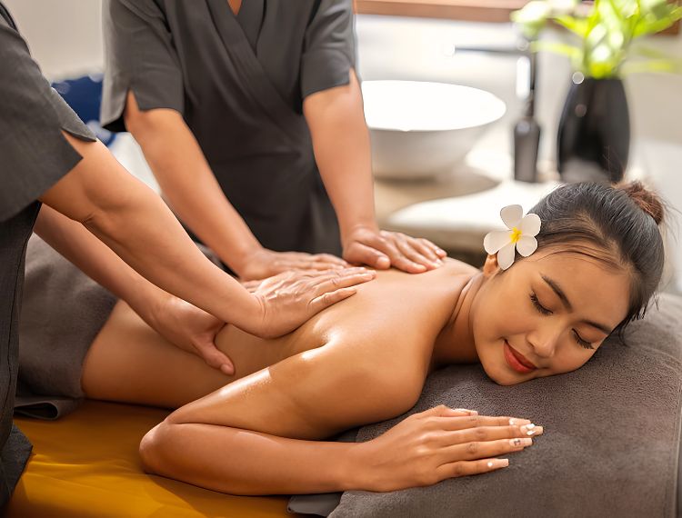 Spa guest receiving a therapeutic two-therapist four-hand back massage with a frangipani flower in her hair.