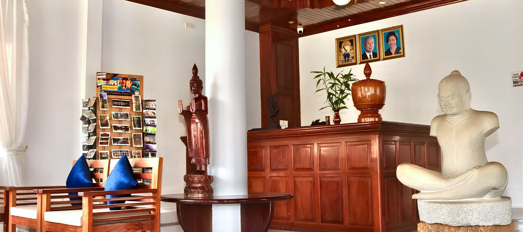 Hotel reception lobby featuring a carved wooden front desk, traditional stone statue, and royal portraits.
